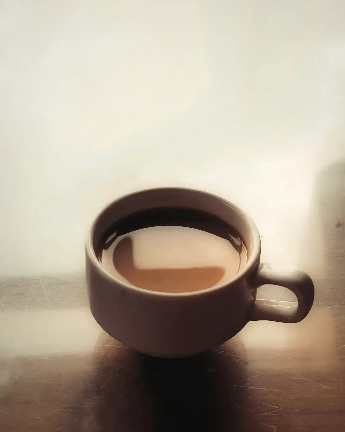 A dark ceramic cup of tea on a reflective table against a bright hazy background